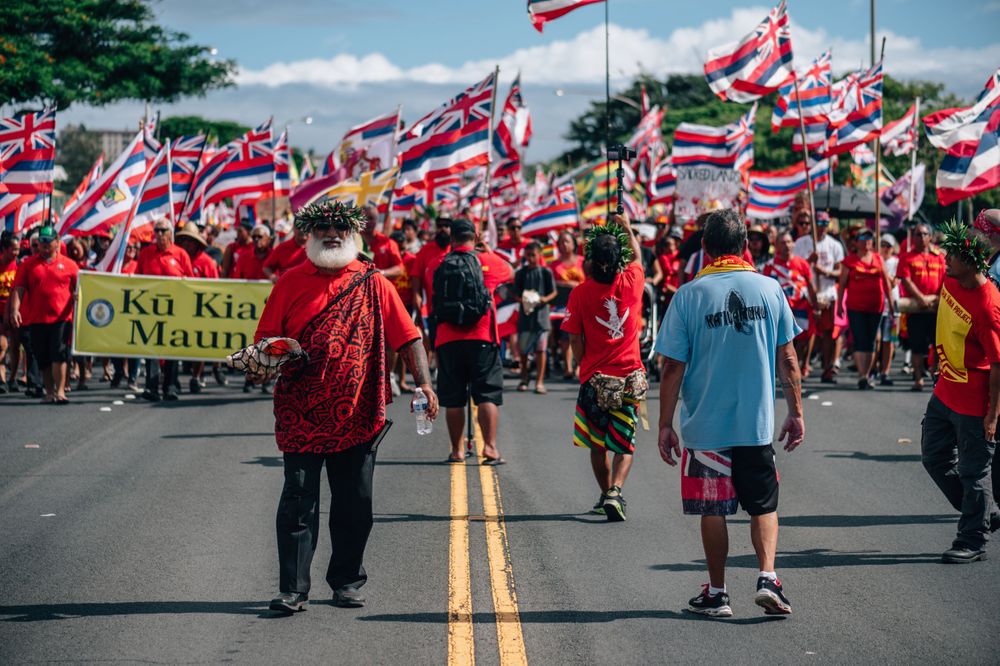 Protesters march down street carrying Hawaiian flags and banners during demonstration.