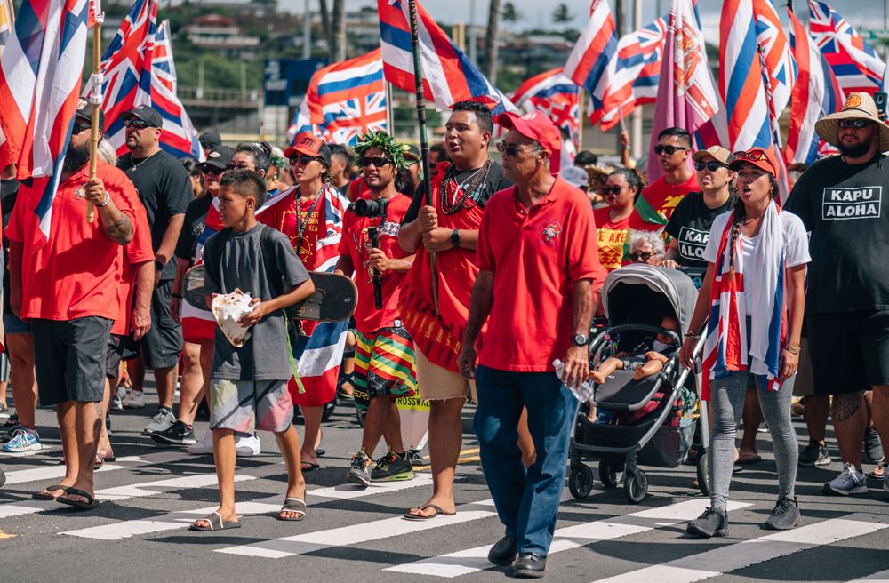 Crowd of demonstrators in red clothing march while carrying Hawaiian state flags.