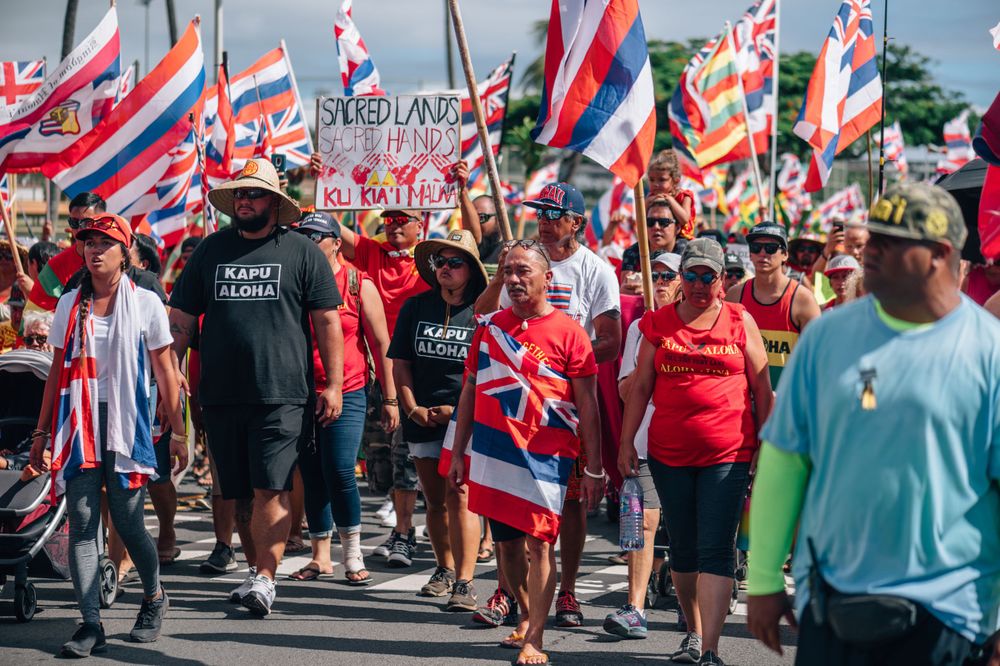 Large group of demonstrators carrying Hawaiian flags march together during street protest.