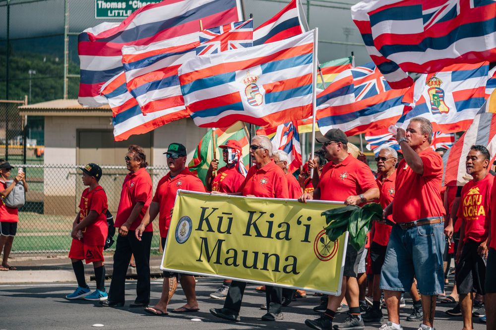 Large crowd of protesters wearing red shirts wave Hawaiian flags during a demonstration.
