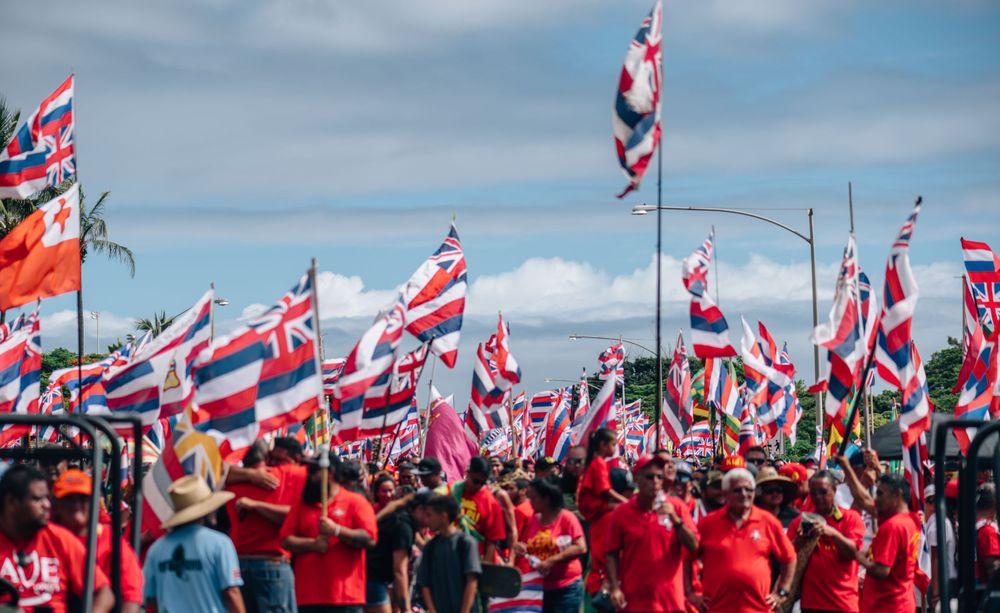 Crowd of protesters in red shirts wave Hawaiian flags against blue sky.