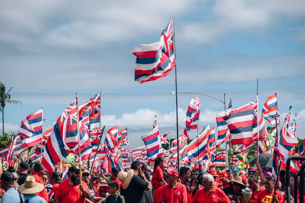 Large protest crowd waves Hawaiian flags under partly cloudy skies.
