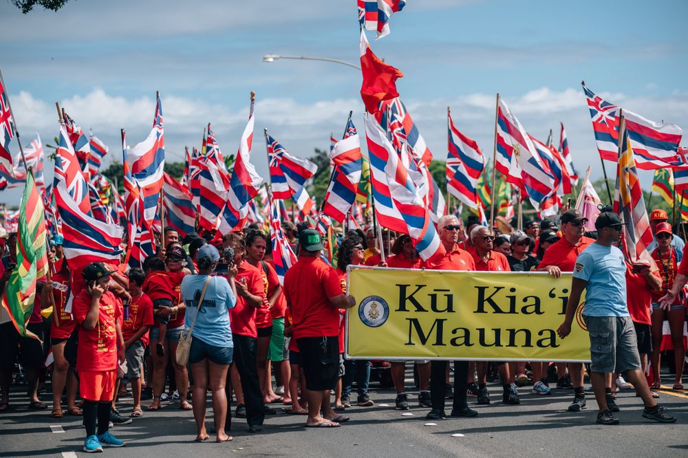 Protesters hold 'Ku Kia'i Mauna' banner while waving Hawaiian flags during demonstration.