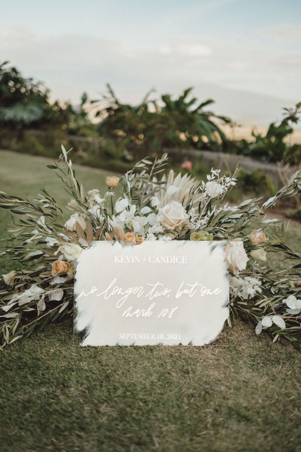 An elegant wedding sign surrounded by white floral arrangements and greenery against a tropical backdrop.
