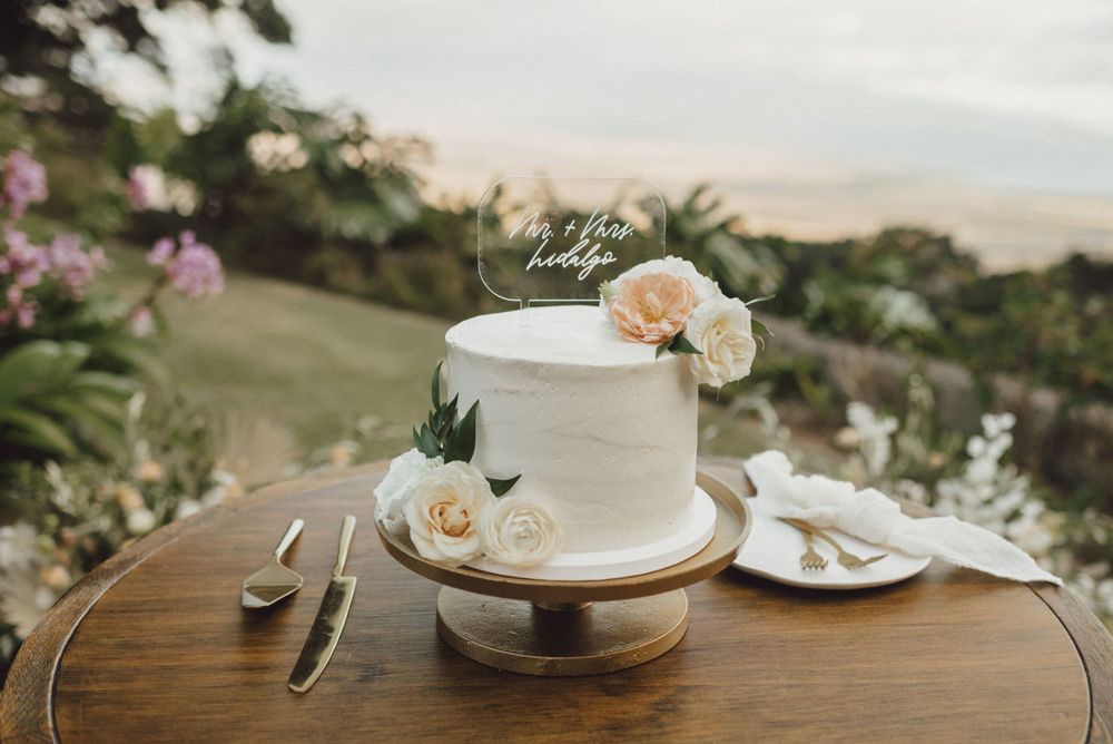 Elegant white wedding cake with peach floral decorations sits on rustic wooden table at outdoor venue.