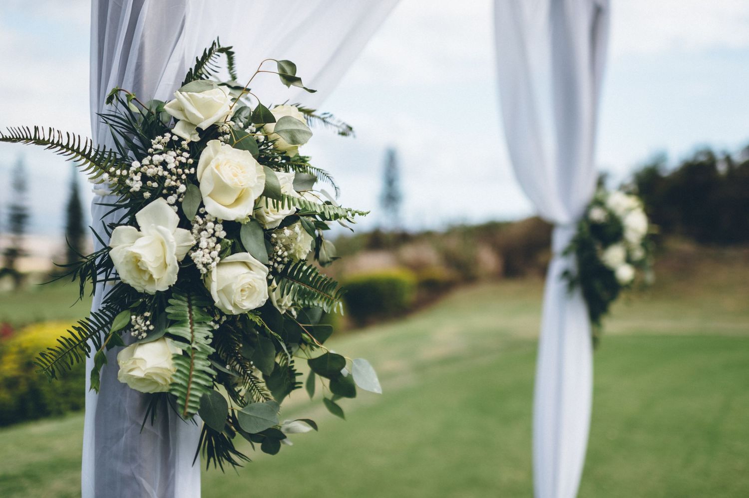 White roses and greenery floral arrangement decorates wedding arch draped with white fabric.