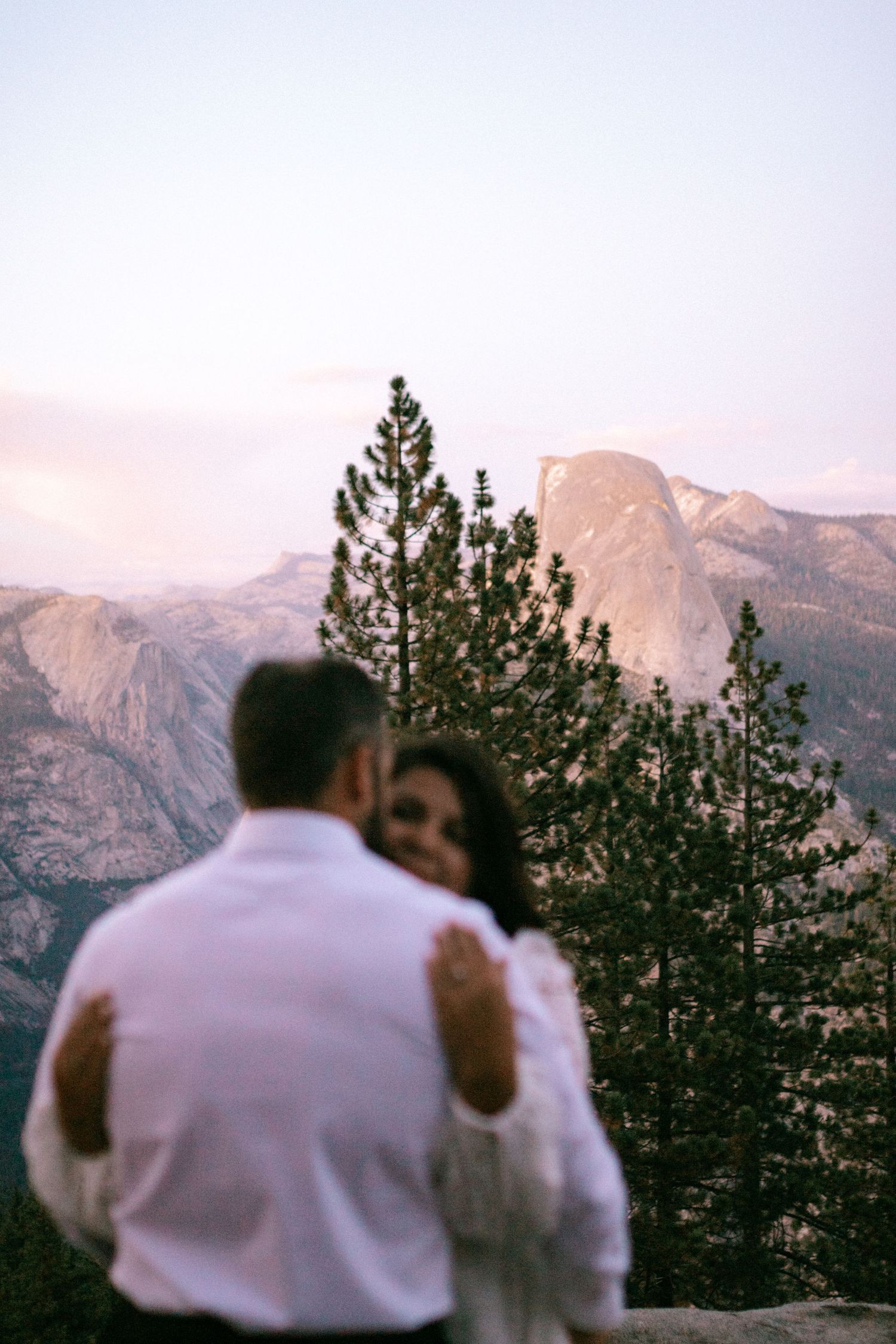 A couple in white embraces while looking at a mountain landscape with evergreen trees at sunset.