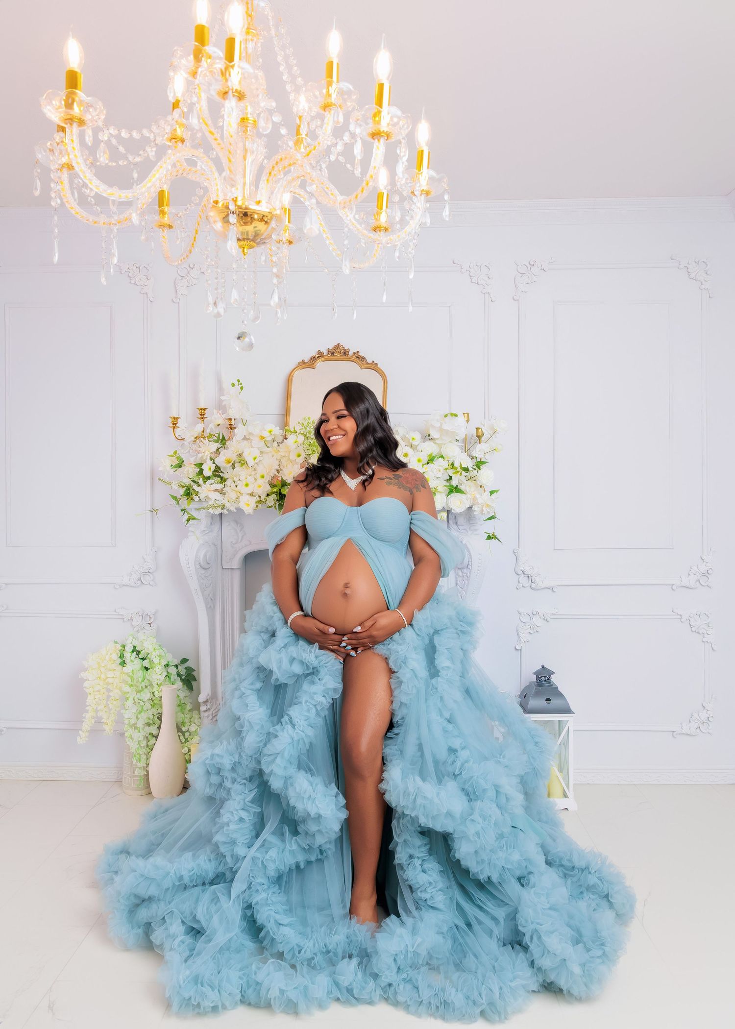 Pregnant woman in a stunning blue tulle gown poses under a crystal chandelier in an elegant white room.
