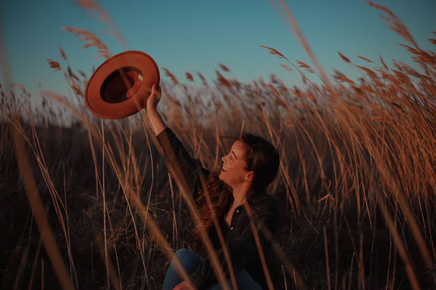Person in white dress and brown hat sitting in sunlit meadow with tall grass during golden hour.