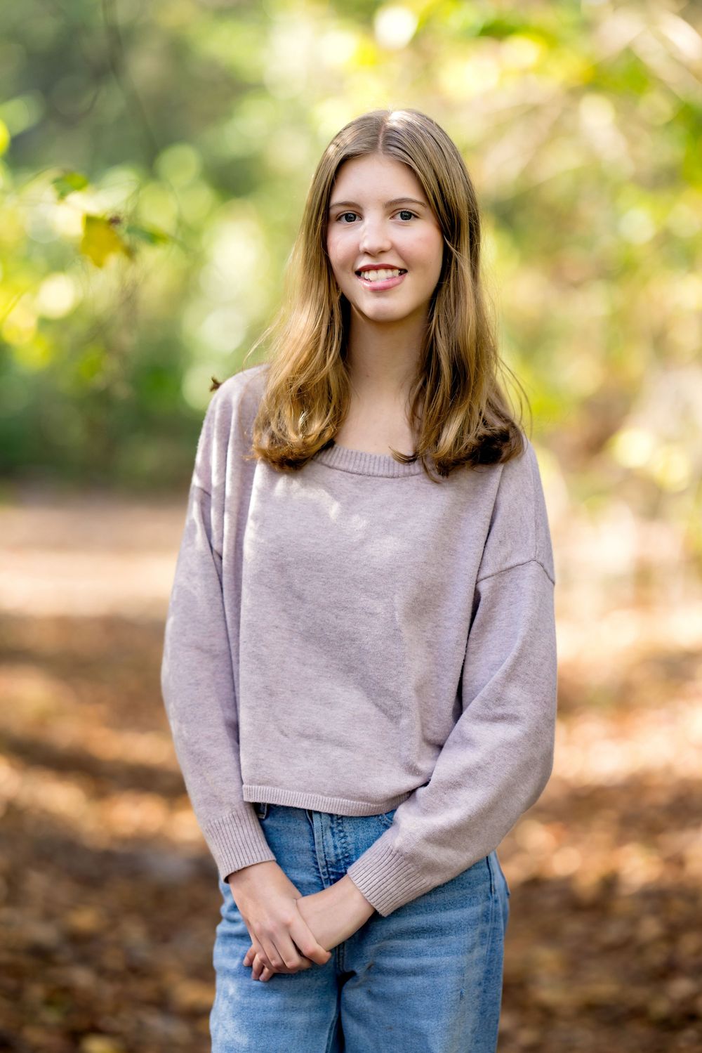 Young person in a casual beige sweater and jeans smiling outdoors with autumn foliage in the background.