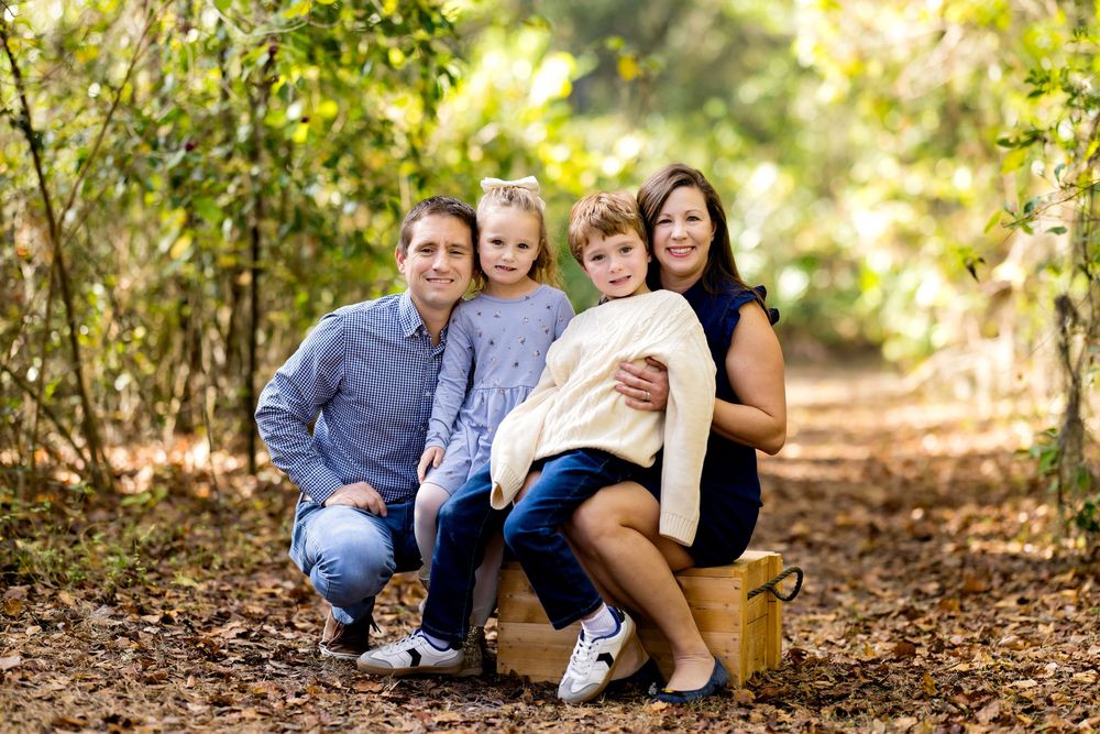 A family of four poses together on a wooded path with sunlight filtering through the trees in the background.