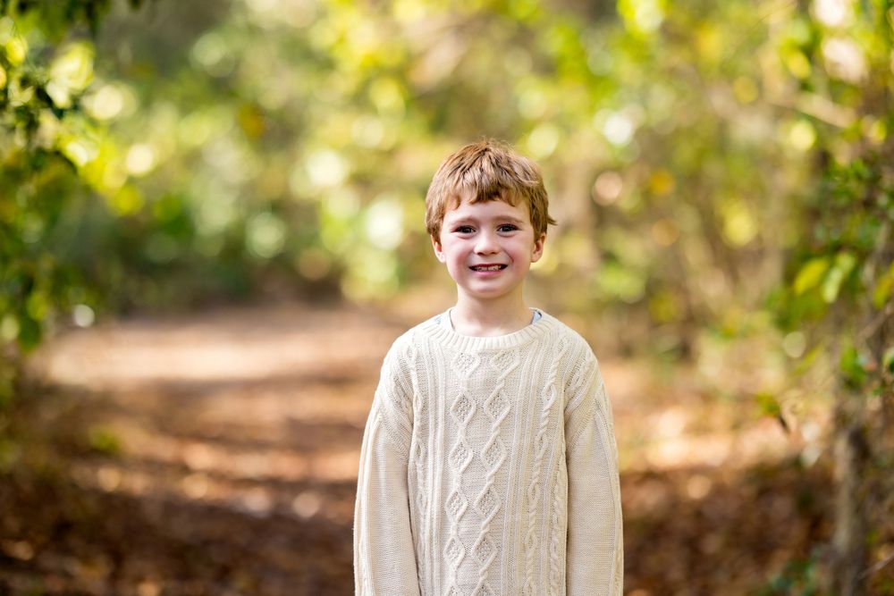 A young person in a cream knit sweater smiles warmly while standing outdoors in front of autumn foliage.
