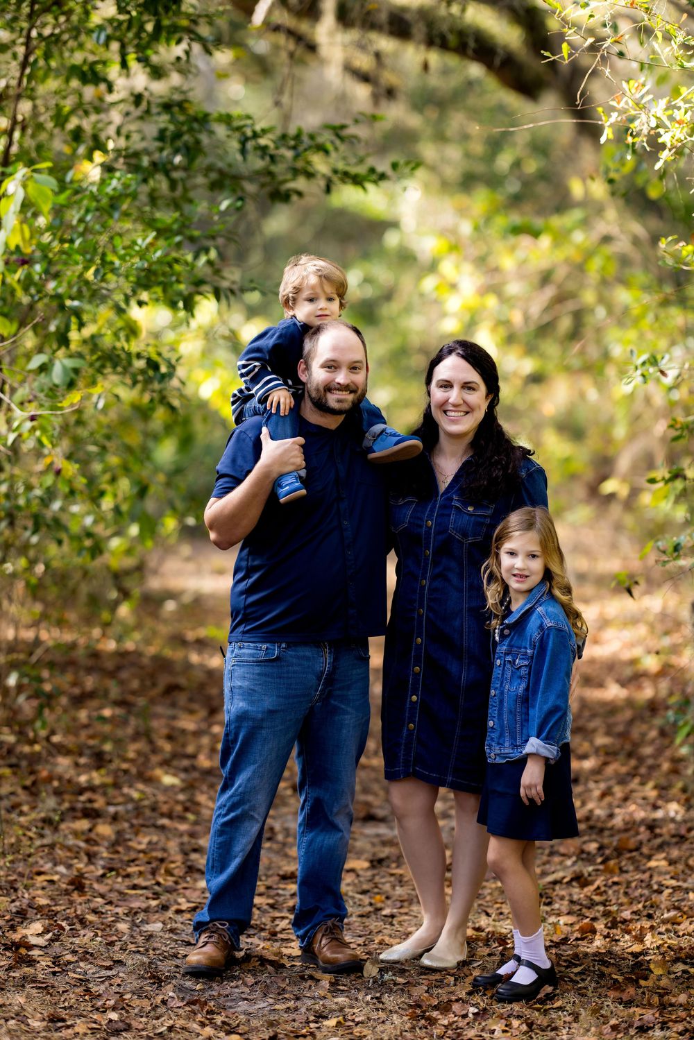 A family of four wearing coordinated navy and denim outfits poses together on a leaf-covered path in an autumn setting.