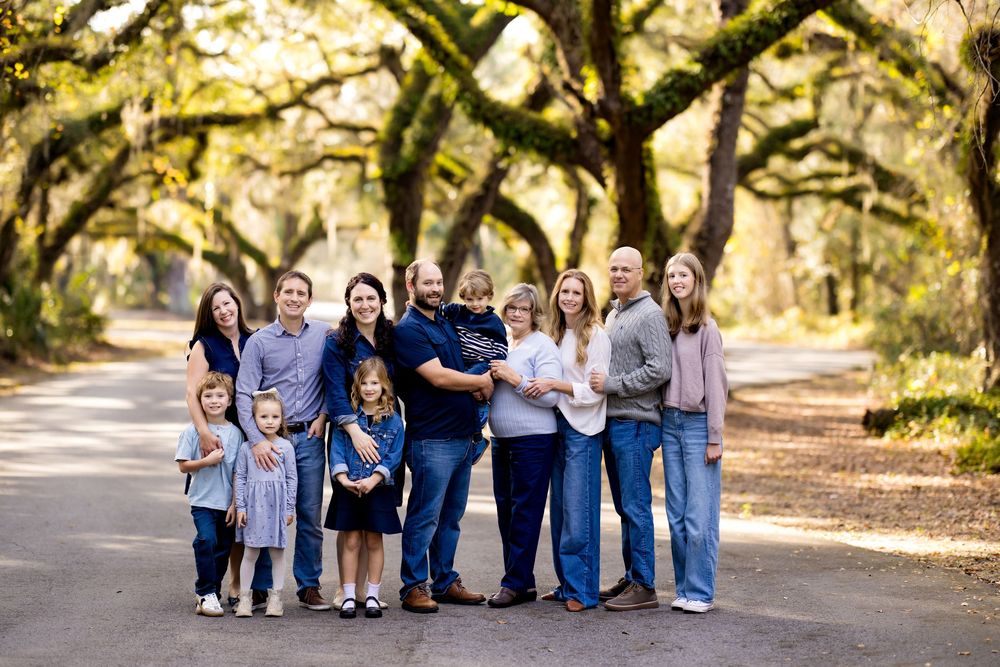 A group in coordinated blue and white outfits poses together on a sunlit tree-lined path during an outdoor photo session.