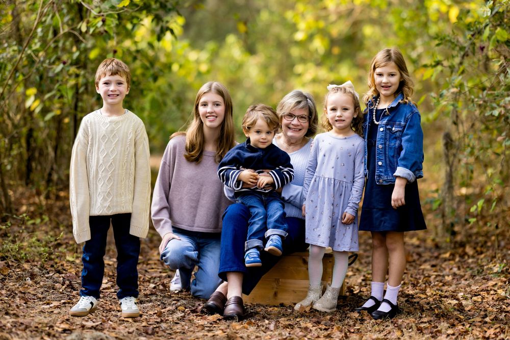 A group sits together on fallen autumn leaves in a wooded outdoor setting wearing coordinated casual clothing.