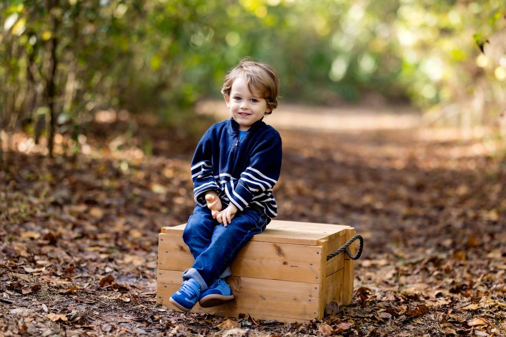 A child sits on a wooden chest in an autumn forest wearing a blue striped sweater and jeans.
