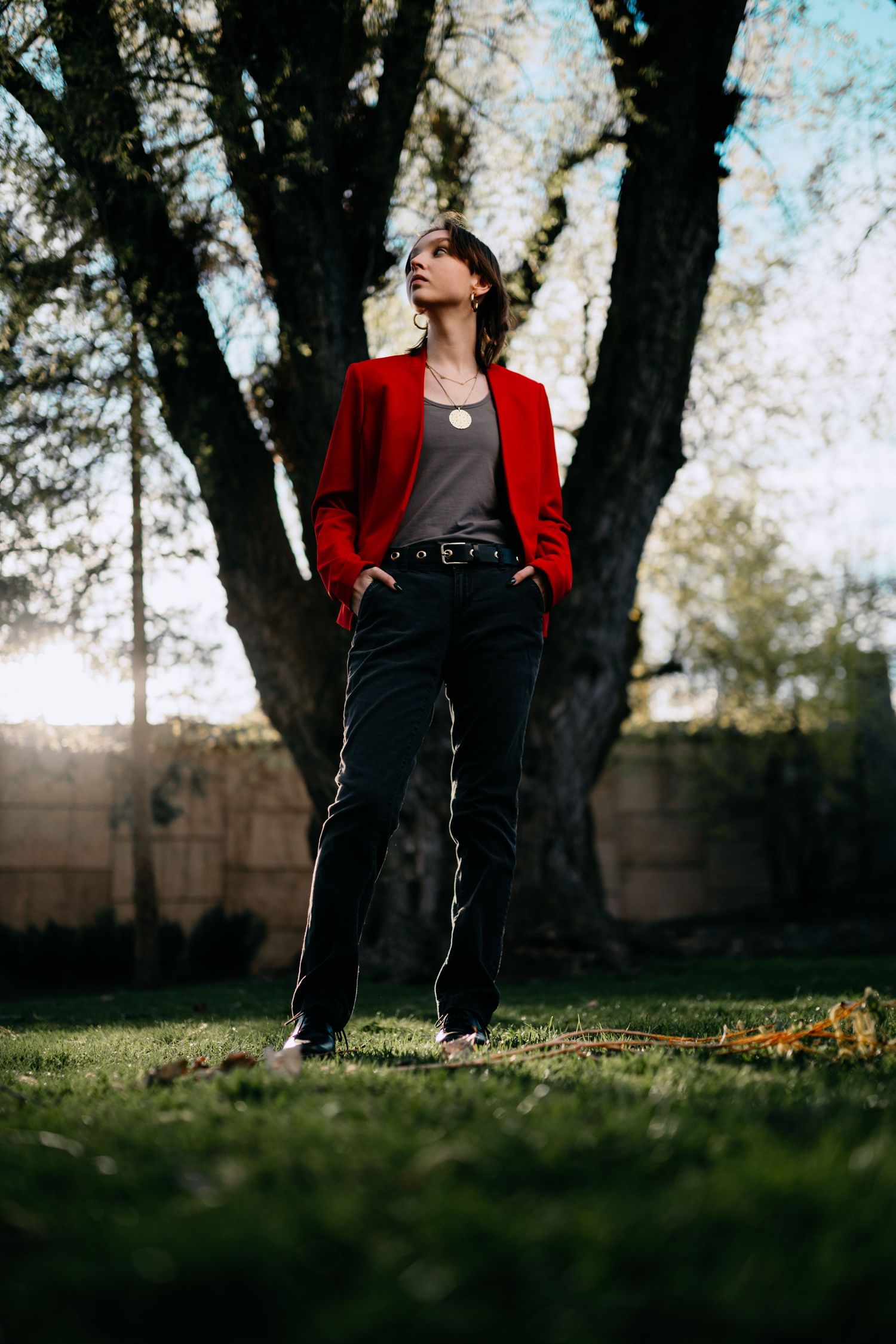 Person in a red blazer and black pants sits thoughtfully against a tree trunk in a grassy outdoor setting.
