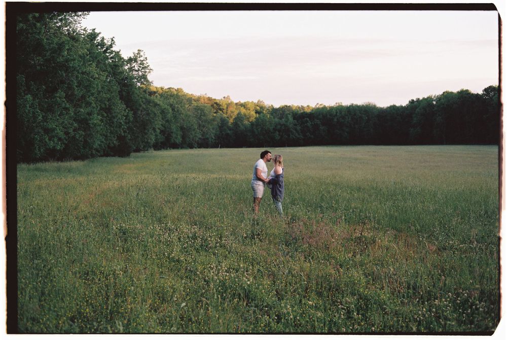 Two figures stand together in a serene grassy meadow at dusk with a tree line visible in the background.