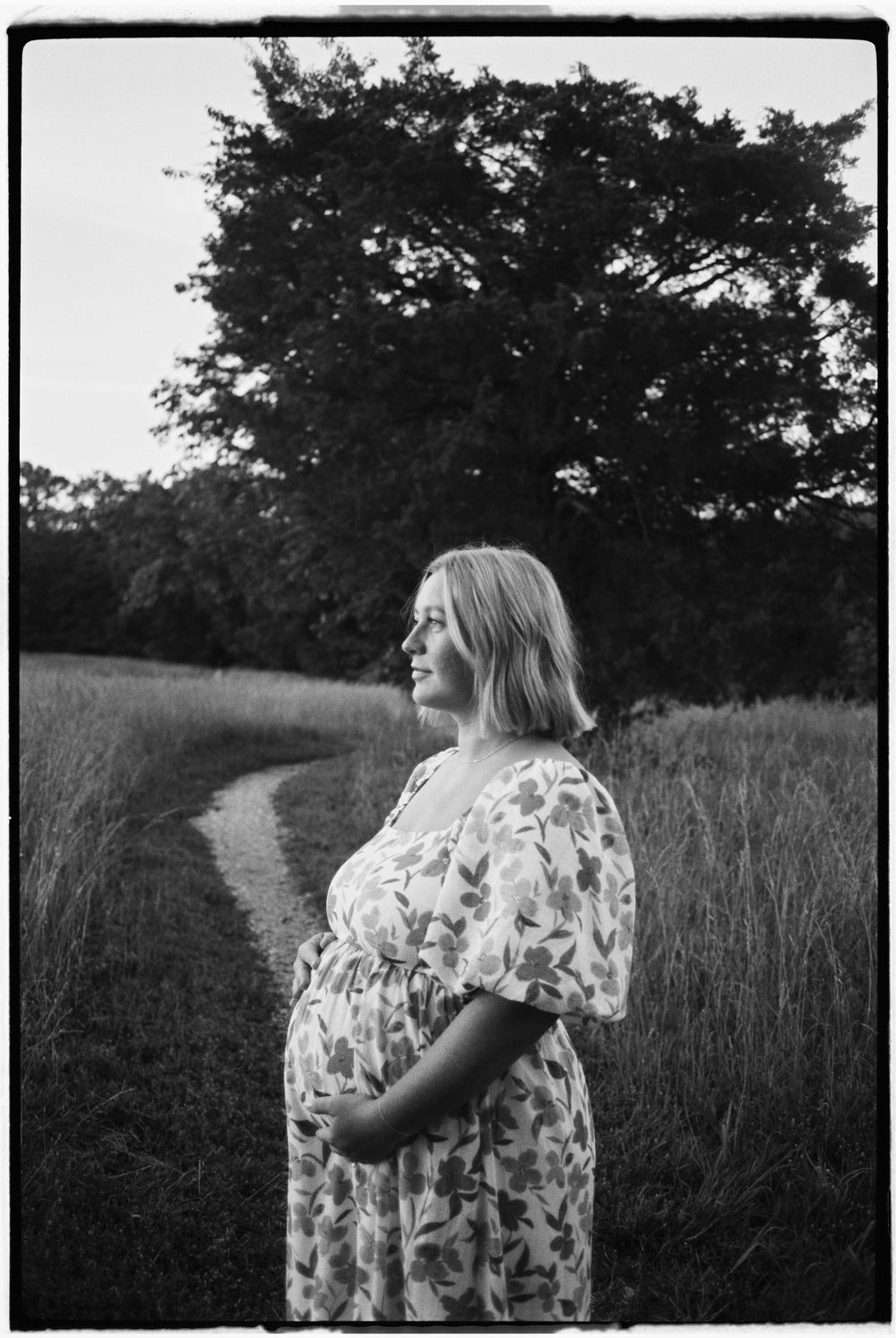 A person in a floral dress walks along a rural path through tall grass at dusk.