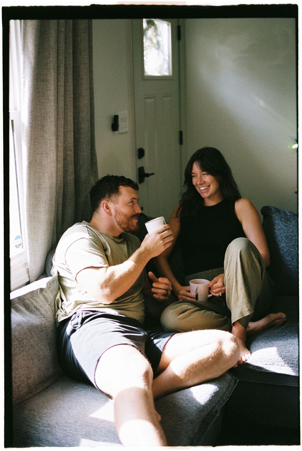 Two people share a relaxed moment together on a couch in natural window light.