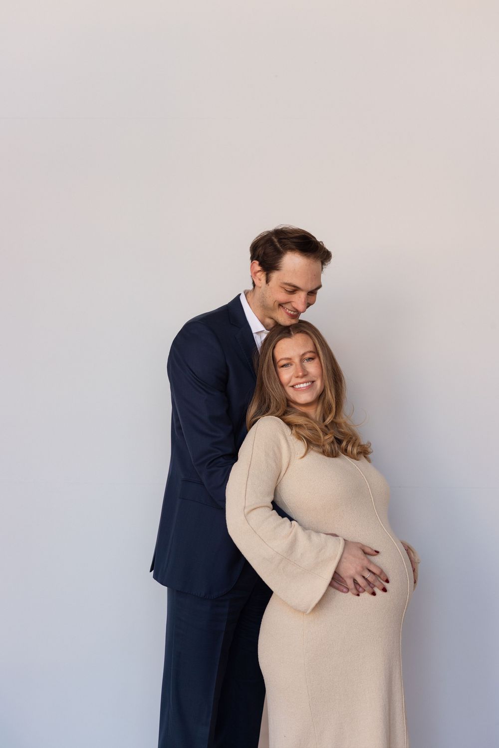 A couple in formal attire embraces during a maternity photo shoot against a light background.
