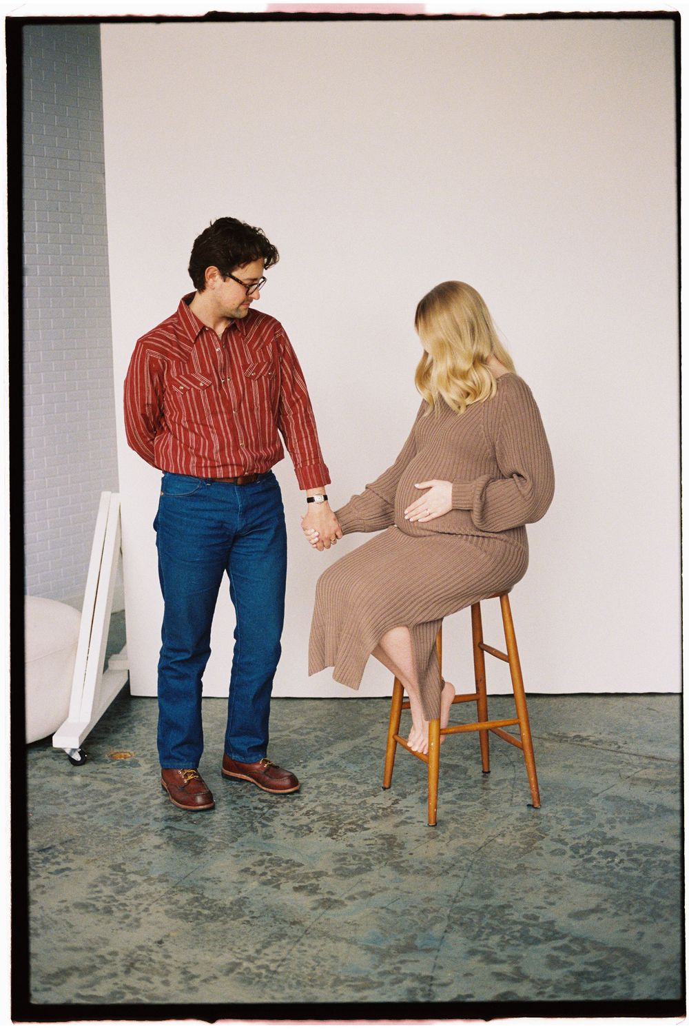 Two people in casual attire interact in a minimalist setting with a wooden stool against a plain wall.