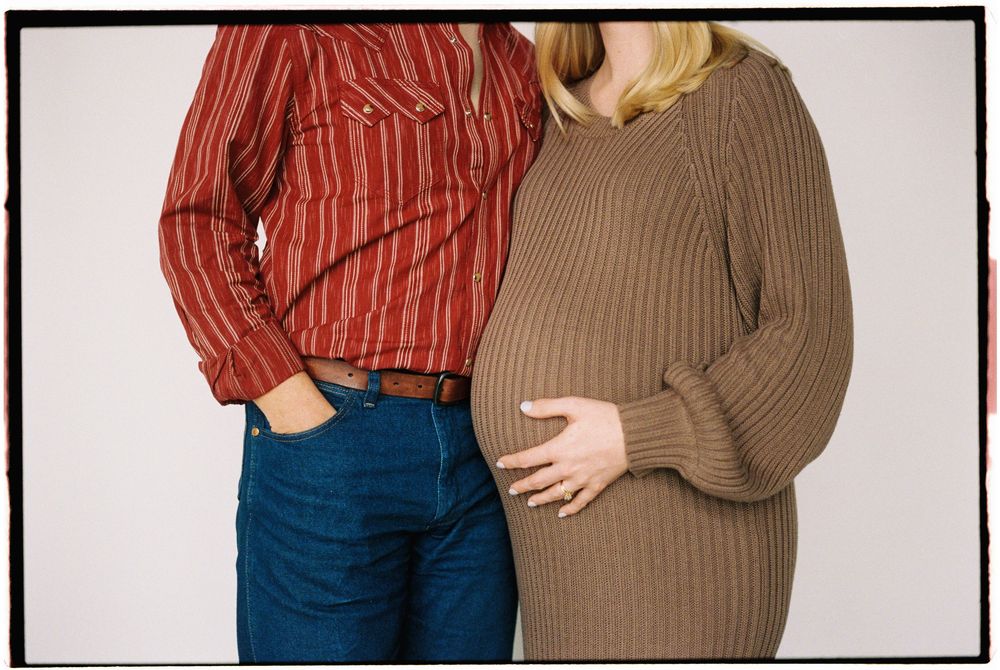 Two people in casual clothing stand close together wearing a striped red shirt and brown sweater.