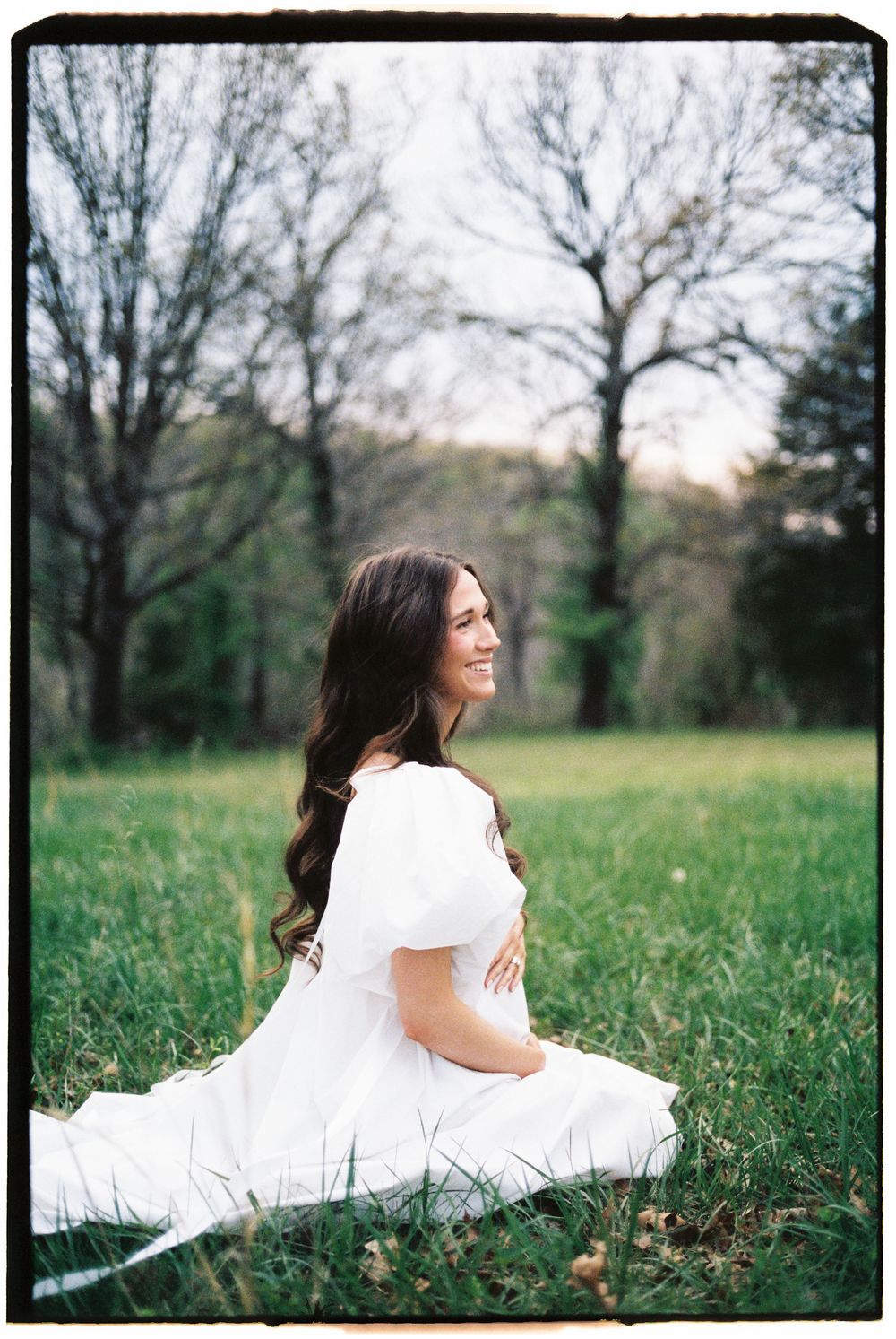 A person in a white dress lying gracefully in a green meadow surrounded by bare trees during springtime.
