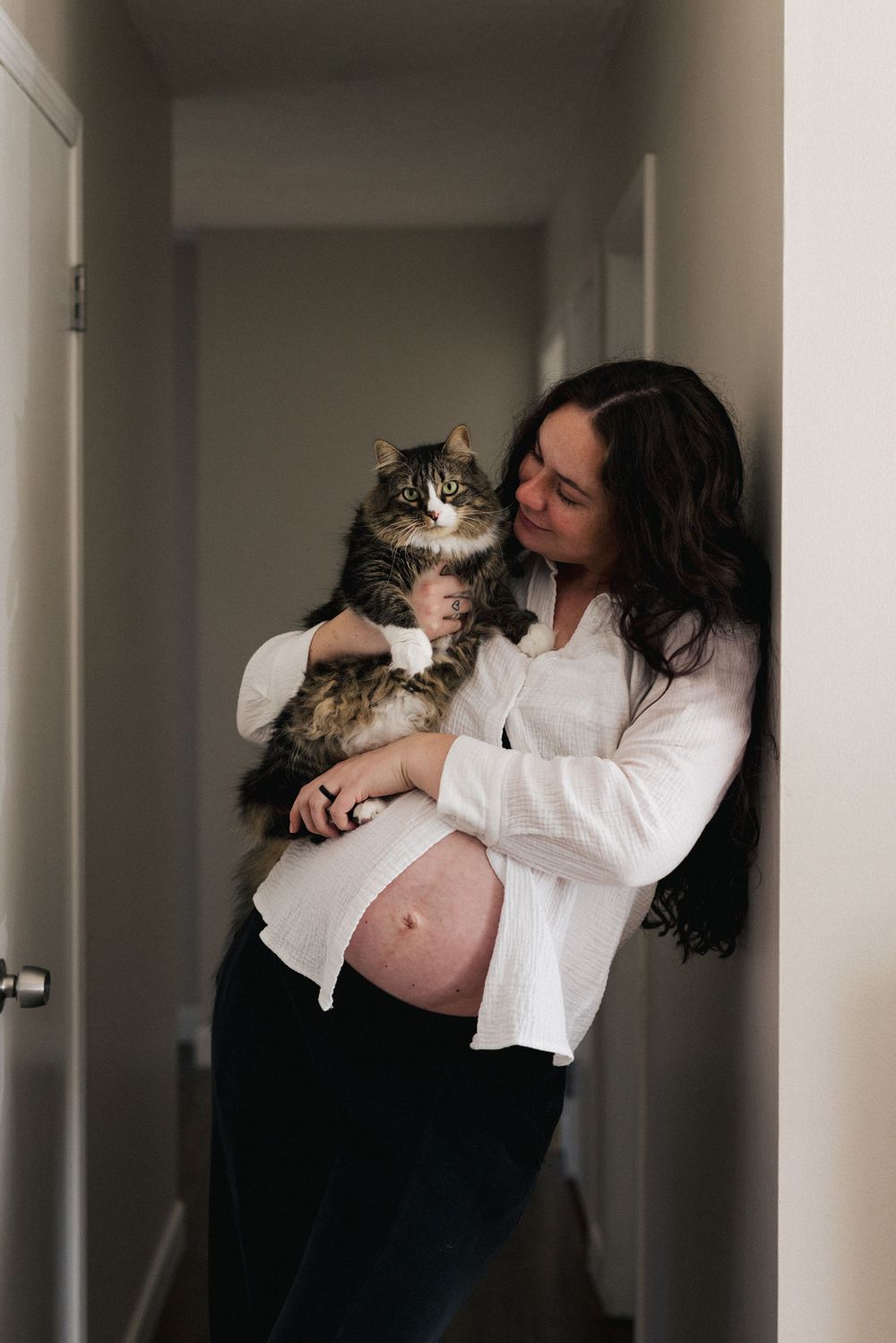 A pregnant woman in a white shirt holds a fluffy Maine Coon cat in a hallway.