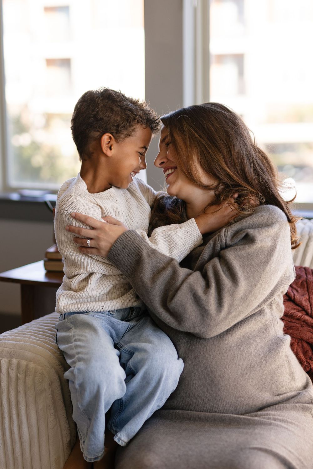 A parent and child share a tender moment of affection while sitting together on a couch in natural lighting.