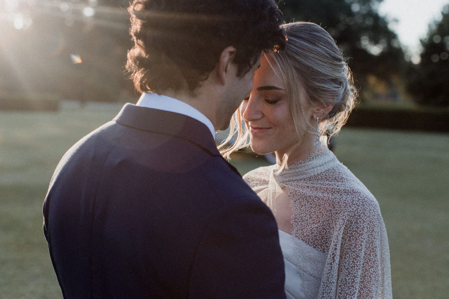 Intimate wedding portrait of newlyweds embracing on manicured lawn during golden hour.