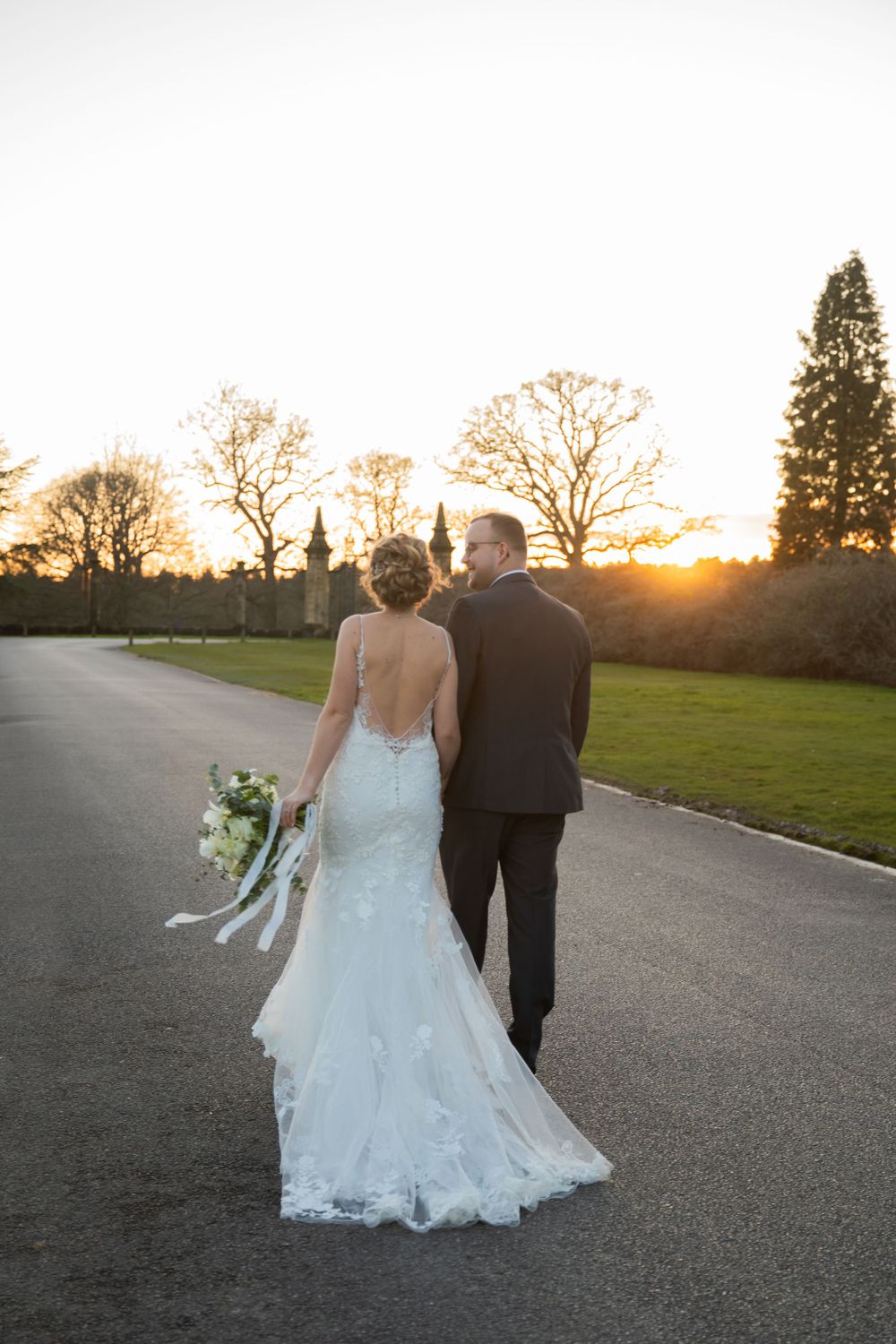 Wedding couple walks hand in hand down path during golden sunset with historic building in background.