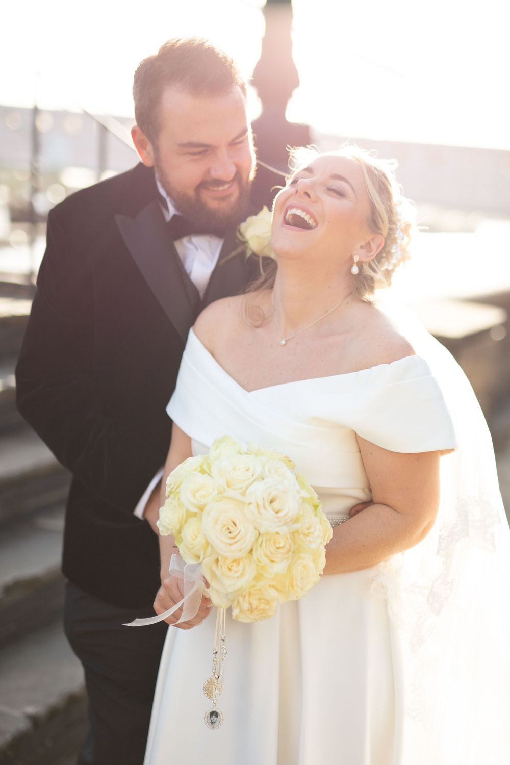 A joyful bride in white holds a bouquet of cream roses while sharing a candid laugh in golden sunlight.