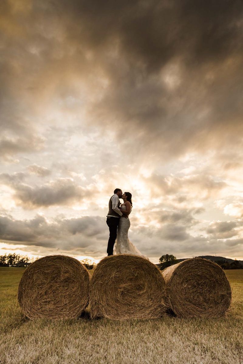 A silhouetted couple stands atop hay bales at sunset with dramatic clouds in the background.