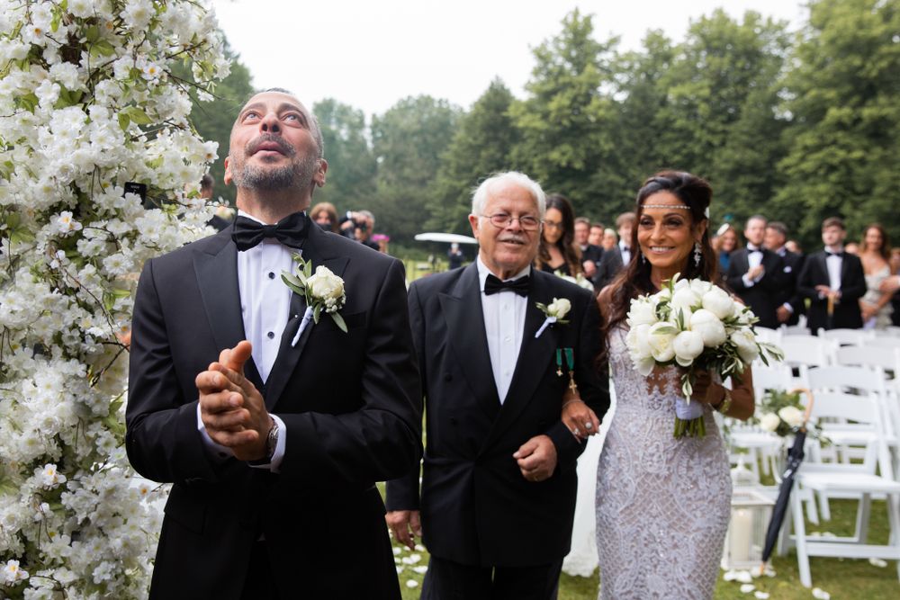Wedding party members in formal attire walk past floral arrangements at outdoor ceremony.