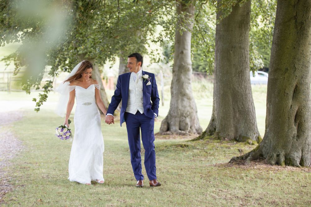 Couple in wedding attire walks hand in hand beneath towering trees.