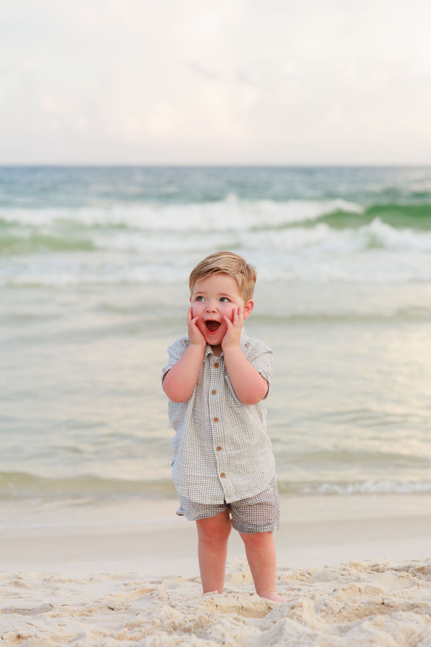 A young child stands on a beach with hands on cheeks expressing excitement as waves roll in behind them.