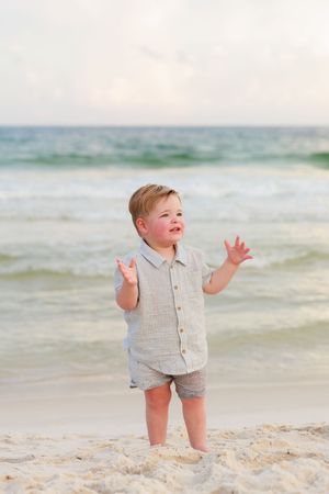 A toddler in beige linen clothing plays joyfully on a sandy beach with ocean waves in the background.