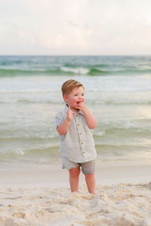 A toddler in a linen shirt and shorts stands joyfully on a sandy beach with ocean waves in the background.