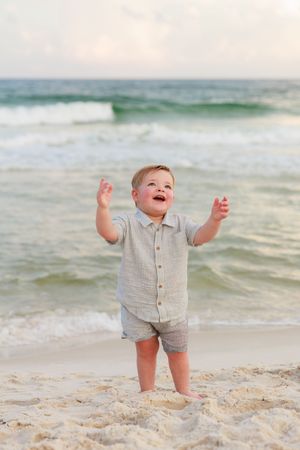 A joyful toddler raises their arms in excitement while standing on a beach with ocean waves in the background.