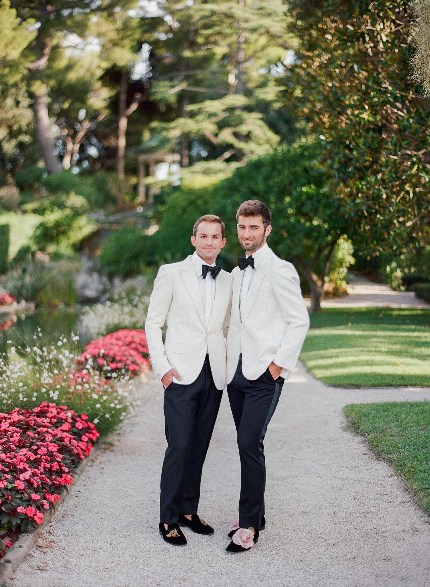 Two people in matching white tuxedo jackets and black pants walk along a garden path lined with pink flowers.