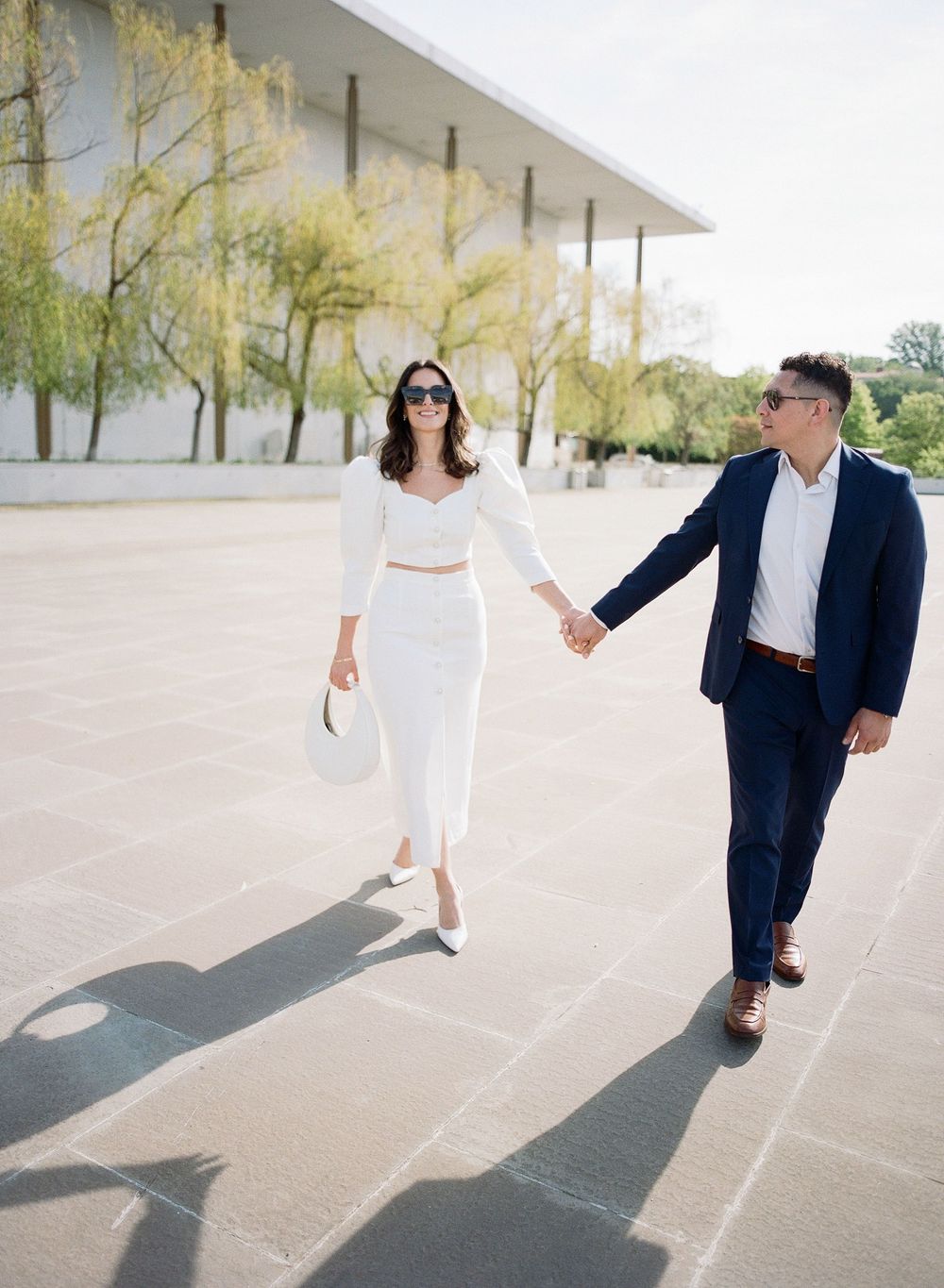 A couple in white and navy outfits playfully dances and poses together in a bright outdoor setting with trees.