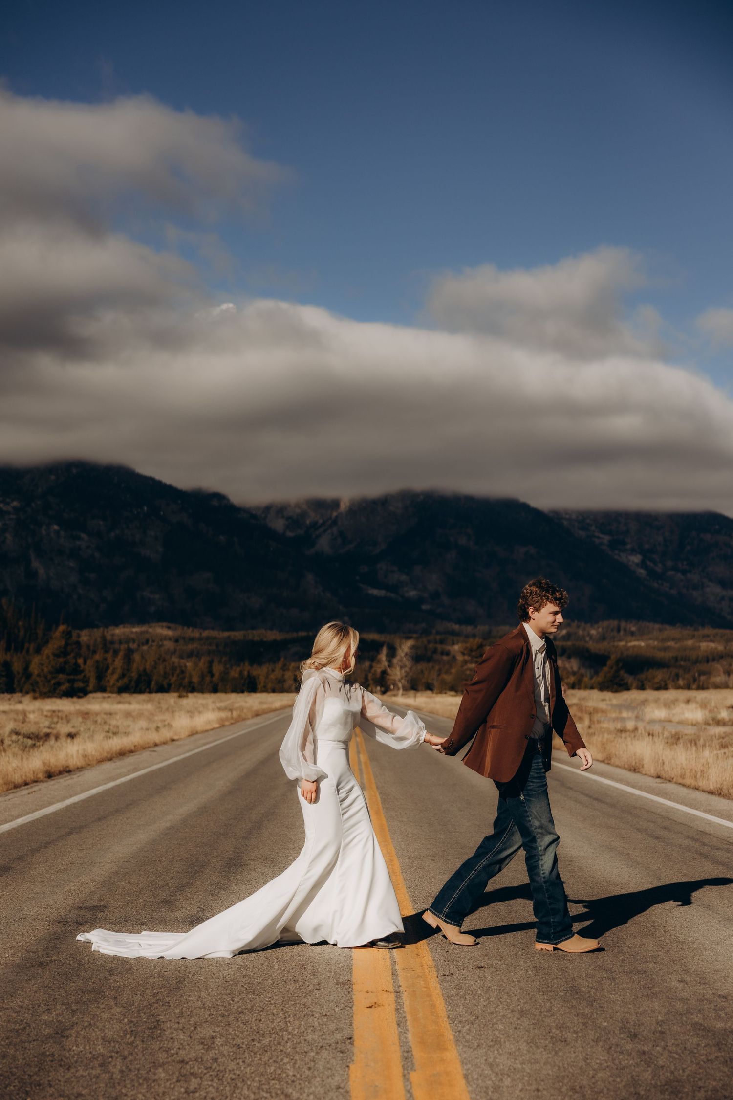 A couple in wedding attire walks and dances along an empty mountain road with dramatic cloudy skies in the background.