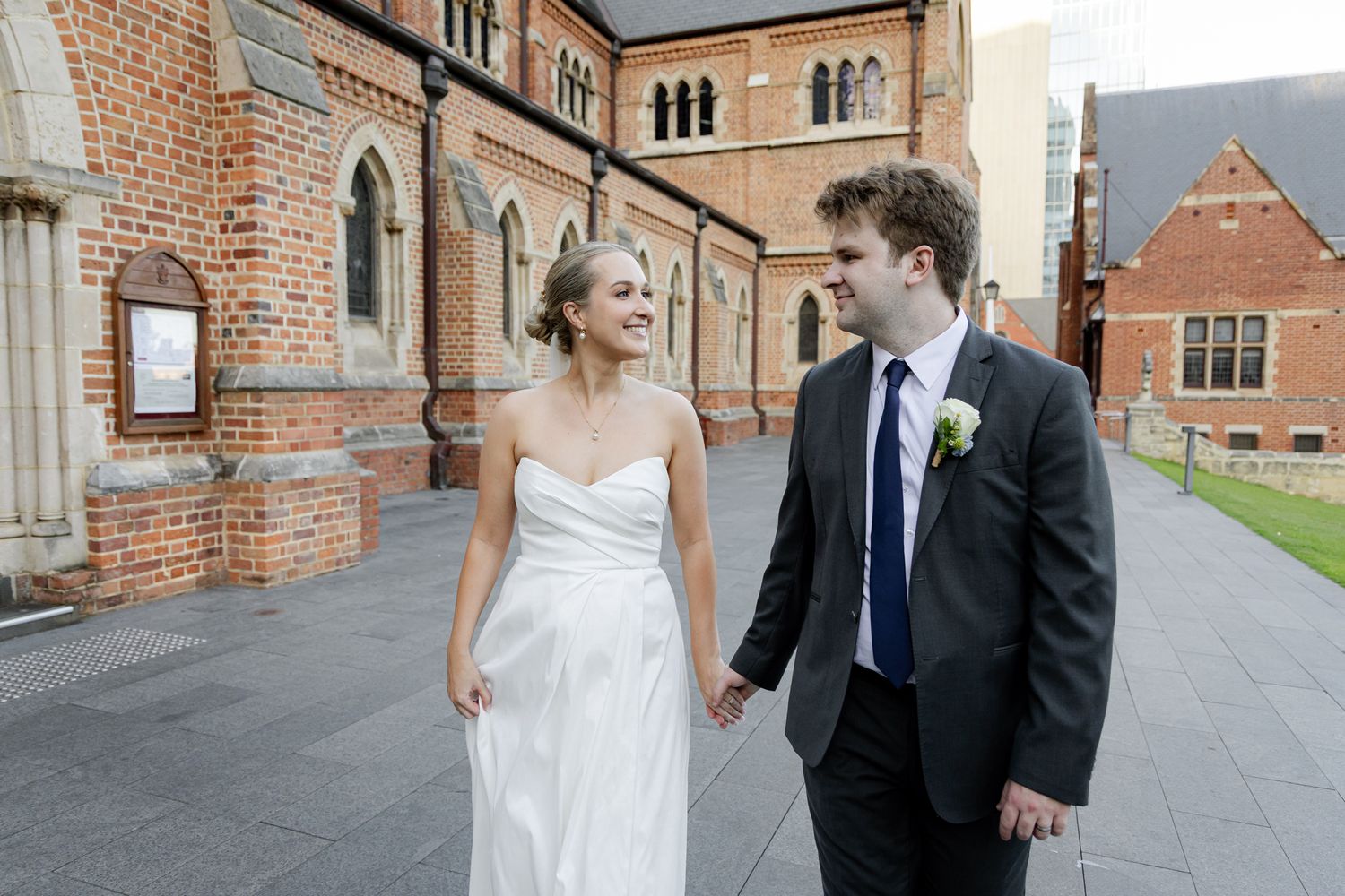 A couple in wedding attire walks together along a brick building exterior while sharing a joyful moment.