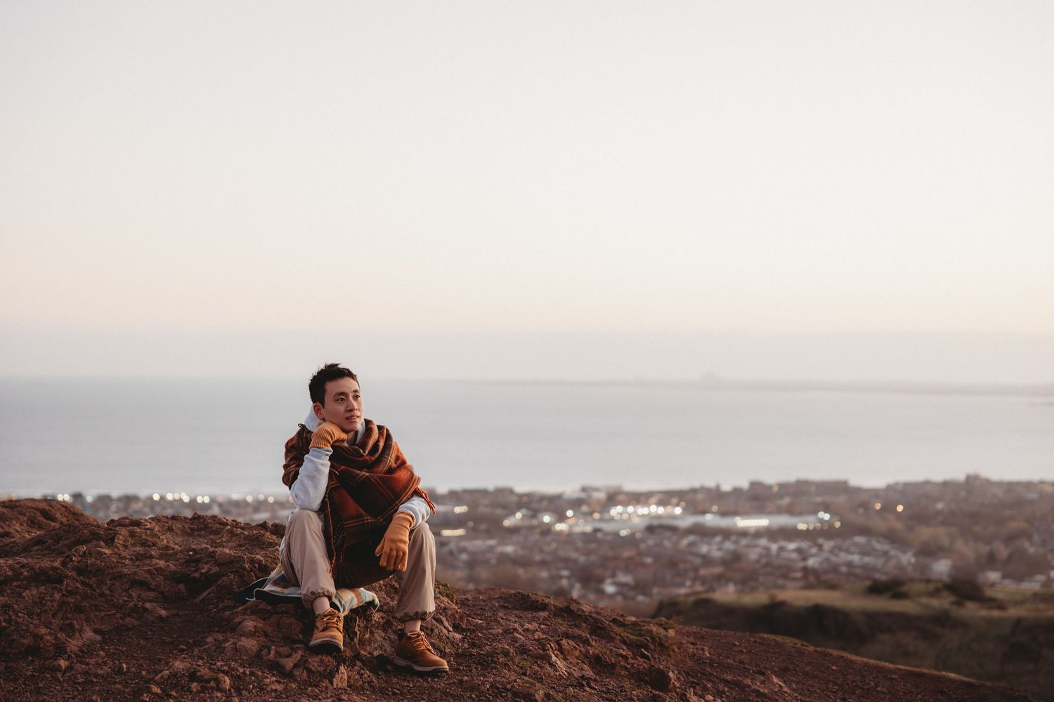 Person sits on rocky overlook at sunset with coastal town and ocean view in the distance.