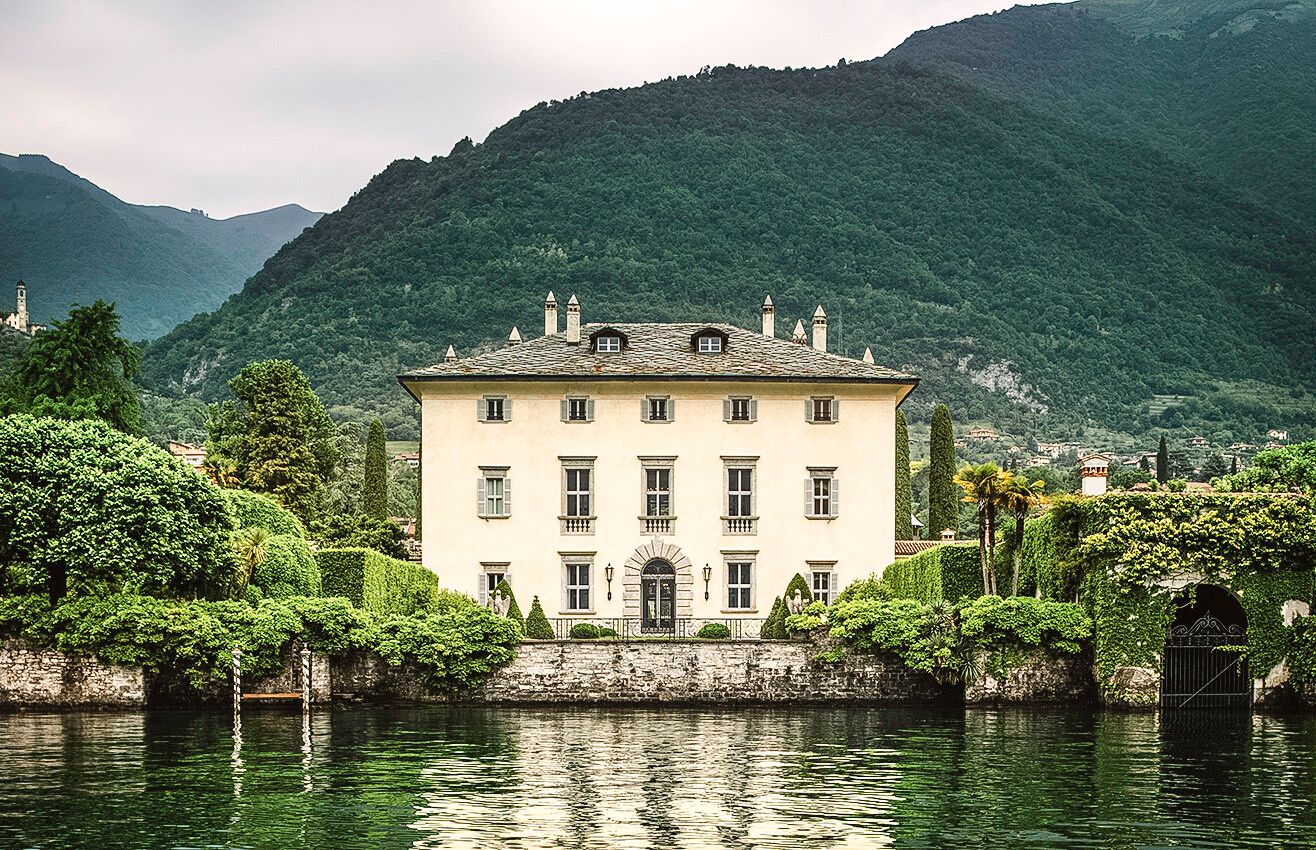 A panoramic view of luxury villas along Lake Como's shoreline with lush green mountains in the background.
