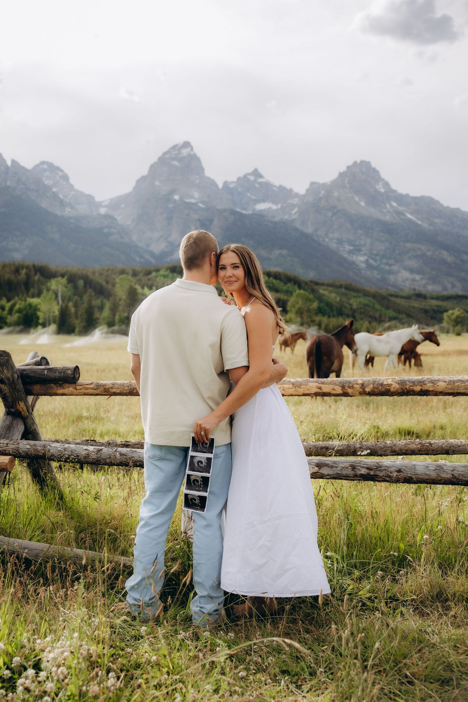 A couple stands by a rustic wooden fence overlooking a meadow with grazing horses and majestic mountain peaks in Wyoming.