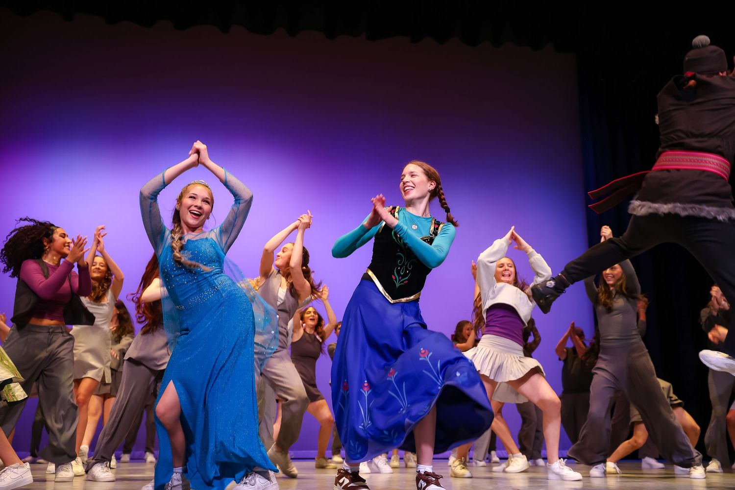 Large group of people wearing white outfits pose together against a bright blue background with a purple circular logo.