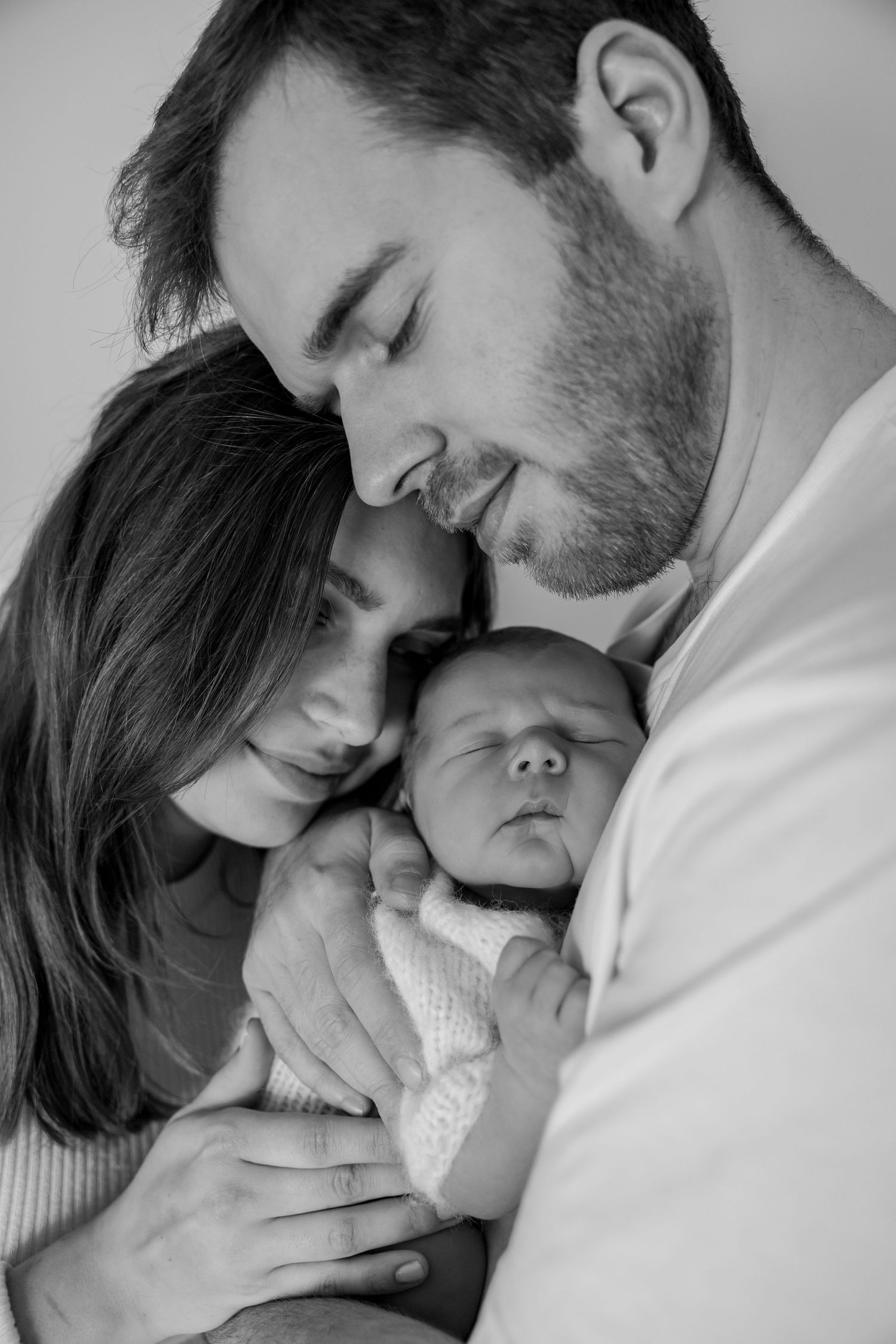 Black and white intimate portrait of parents tenderly embracing their sleeping newborn baby.