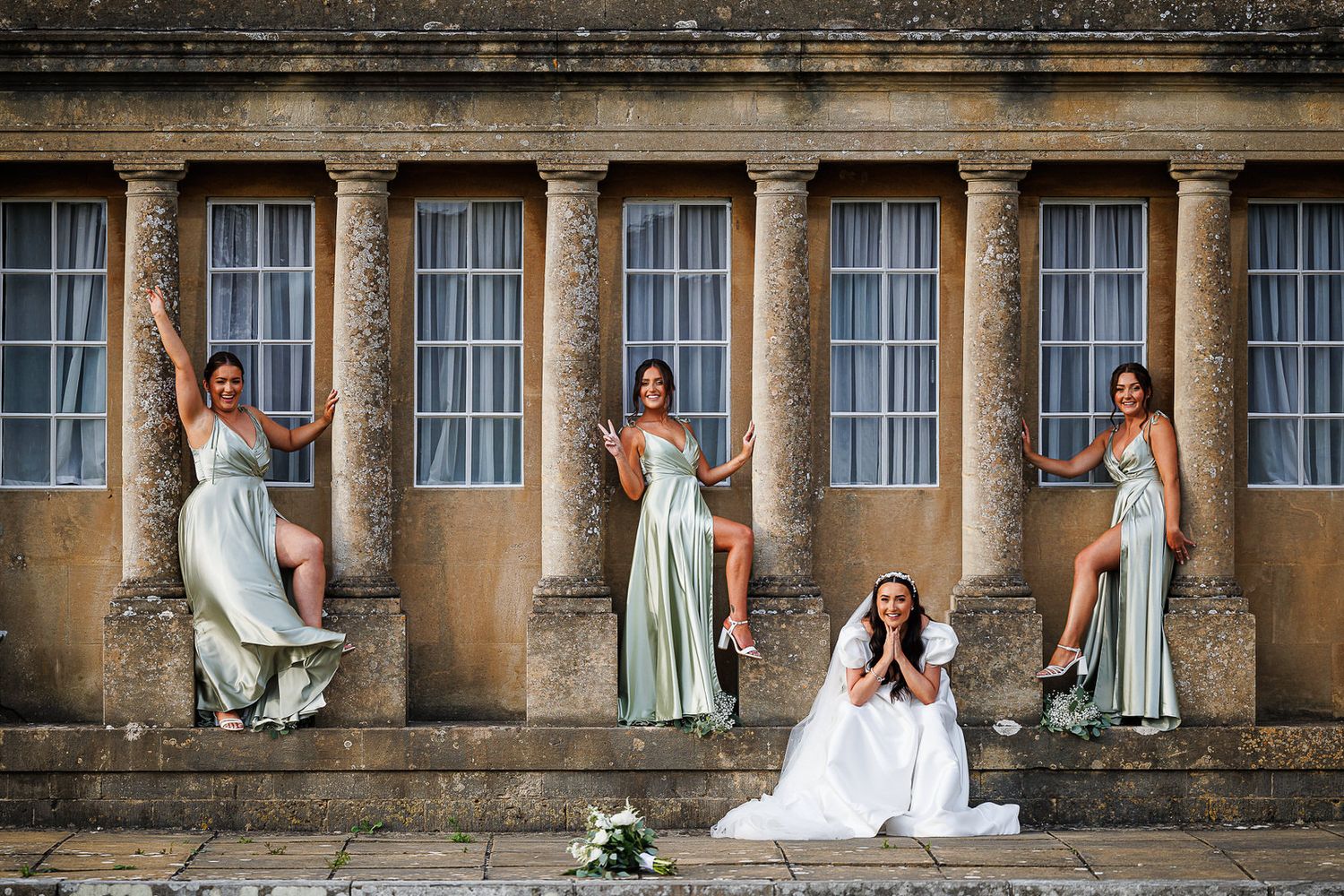 Elegant bridal party in mint green dresses pose gracefully against a historic stone colonnade with tall windows.